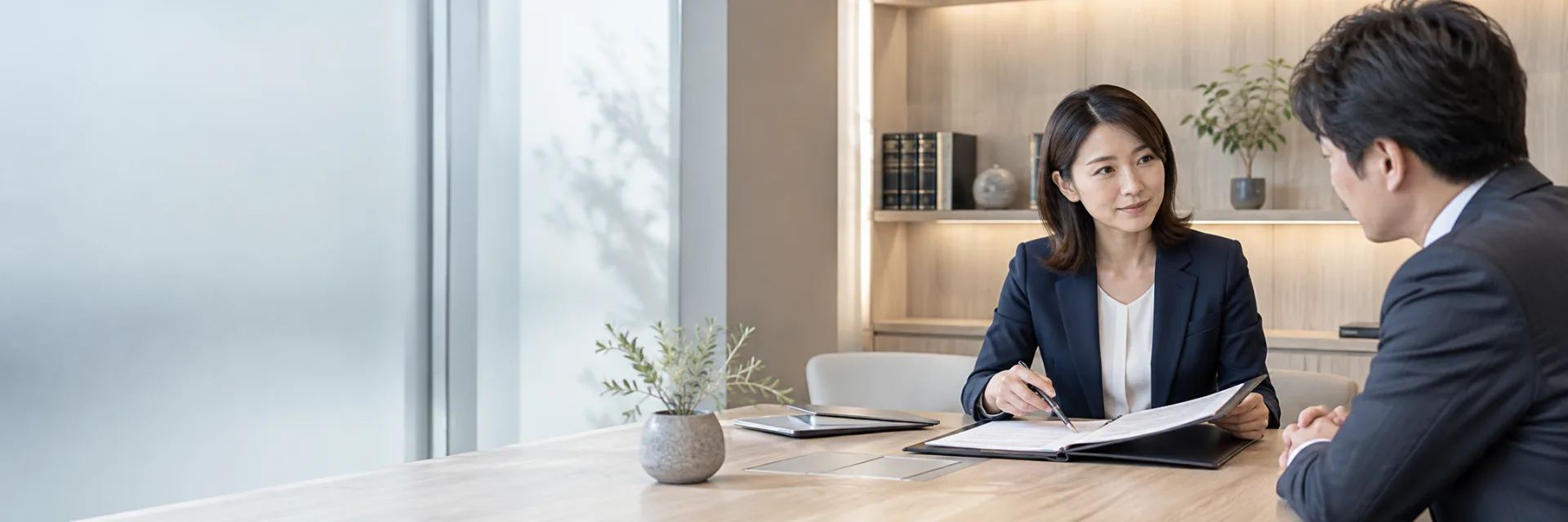 A Japanese female lawyer in a navy suit discussing a neatly organized contract folder with a business client in a refined minimalist law office, discreet oak shelves and frosted glass.